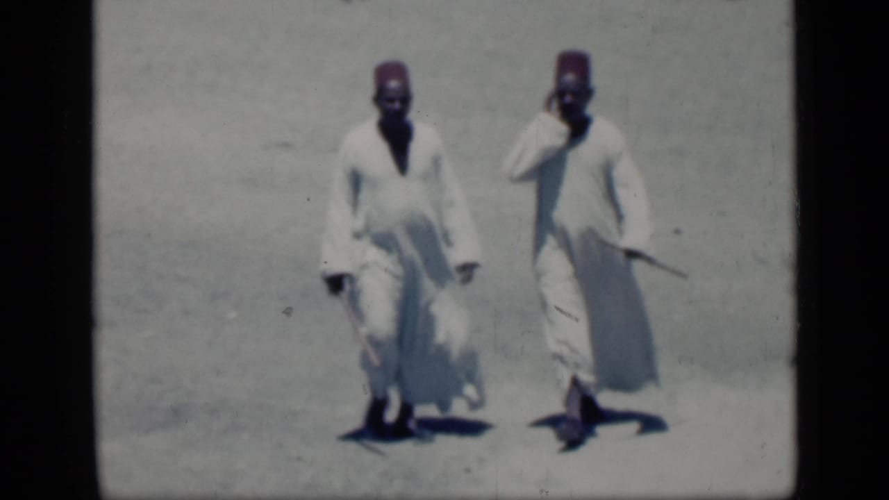 Two robed men in red hats walk across bright open ground carrying staffs.