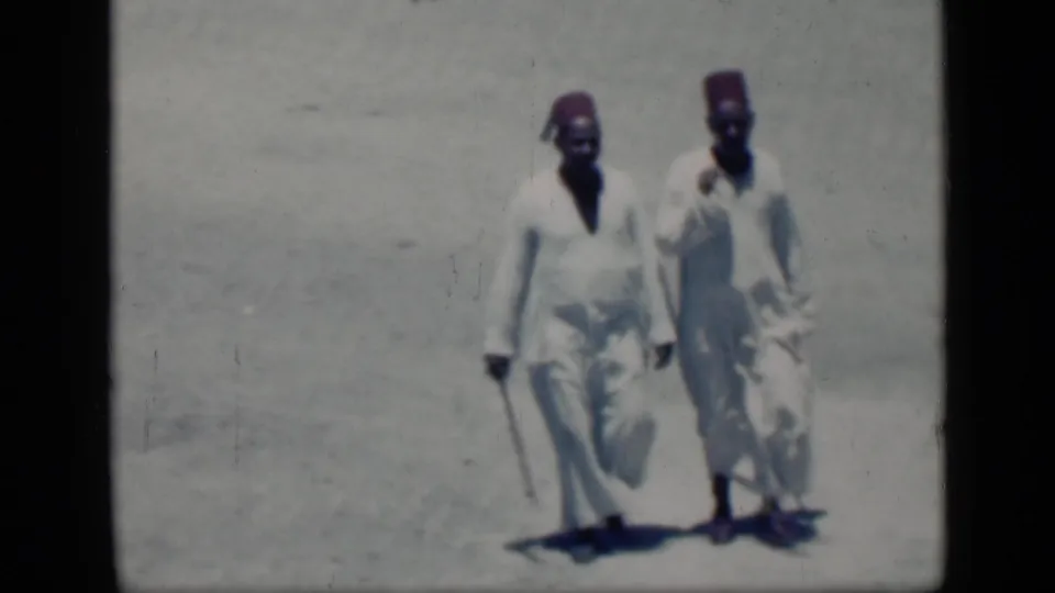 The two men continue walking closer across the bright Cairo ground, their robes and staffs visible in the aged 8mm frame.