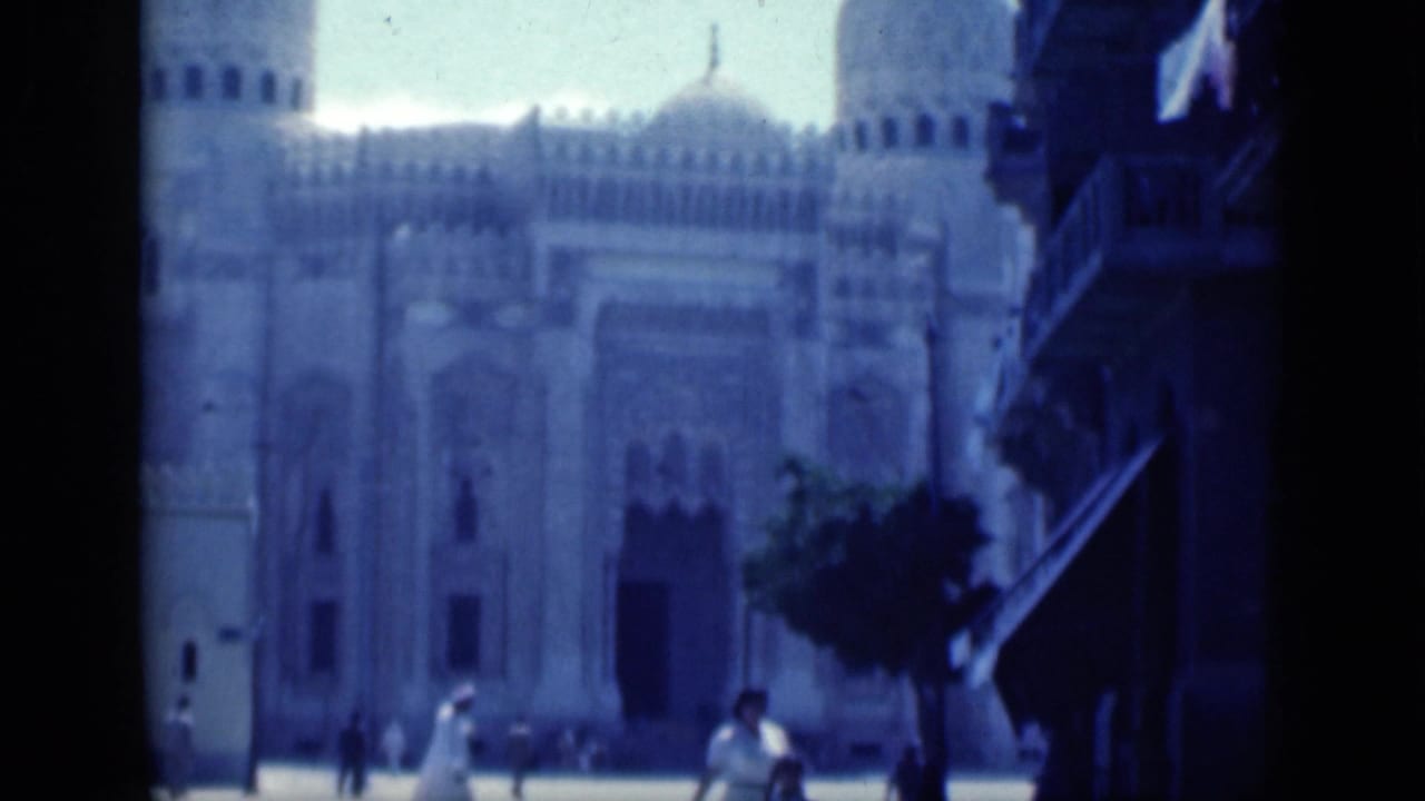 1946 Cairo 8mm frame showing a large domed Islamic landmark across a sunlit street as pedestrians pass in front.