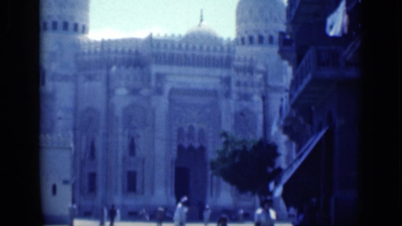 Large mosque exterior with domes, arched details, pedestrians, and shaded street edge.