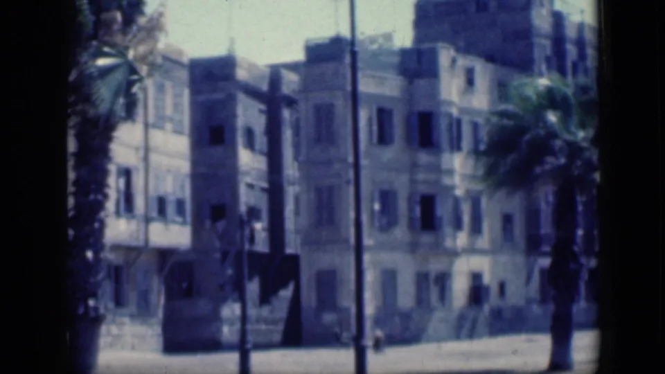 1946 Cairo home-movie frame showing a pale apartment block, palm trunks, street poles, and sunlit pavement.