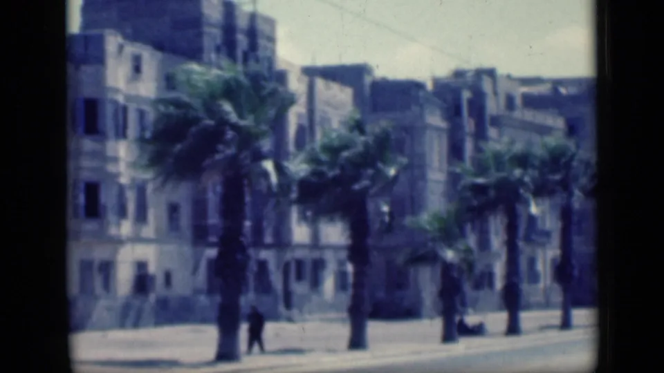 1946 Cairo view with pedestrians near a palm-lined sidewalk, pale buildings, shuttered windows, and a quiet roadway.