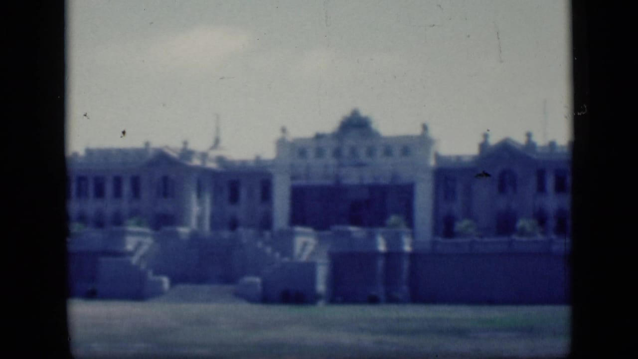 1946 Cairo home-movie frame showing a distant classical-style building facade with arches, windows, terraces, and a blue film cast.