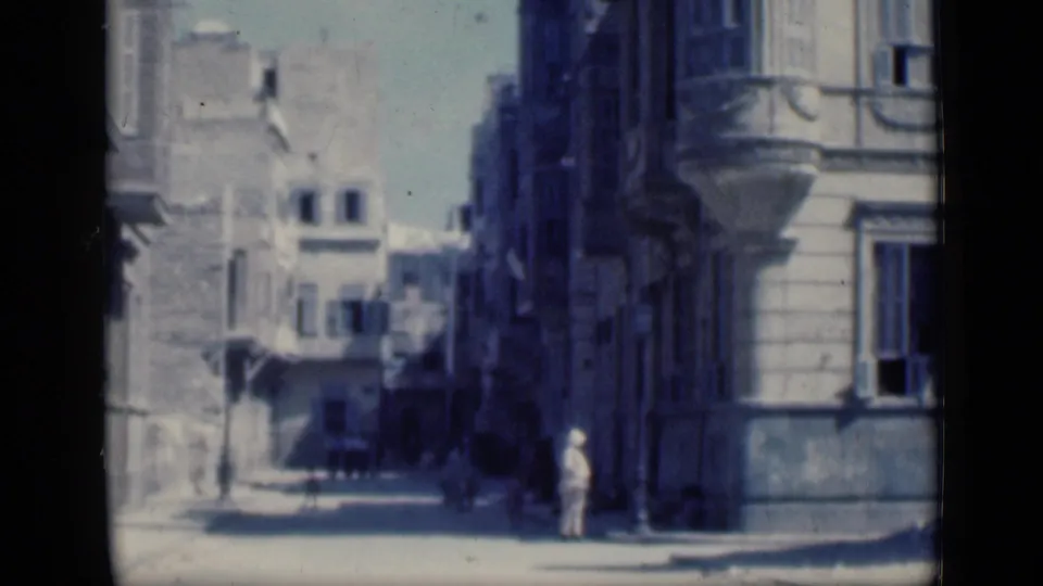 1946 Cairo street corner with a person in light clothing, shadowed building fronts, balconies, and soft blue 8mm color.
