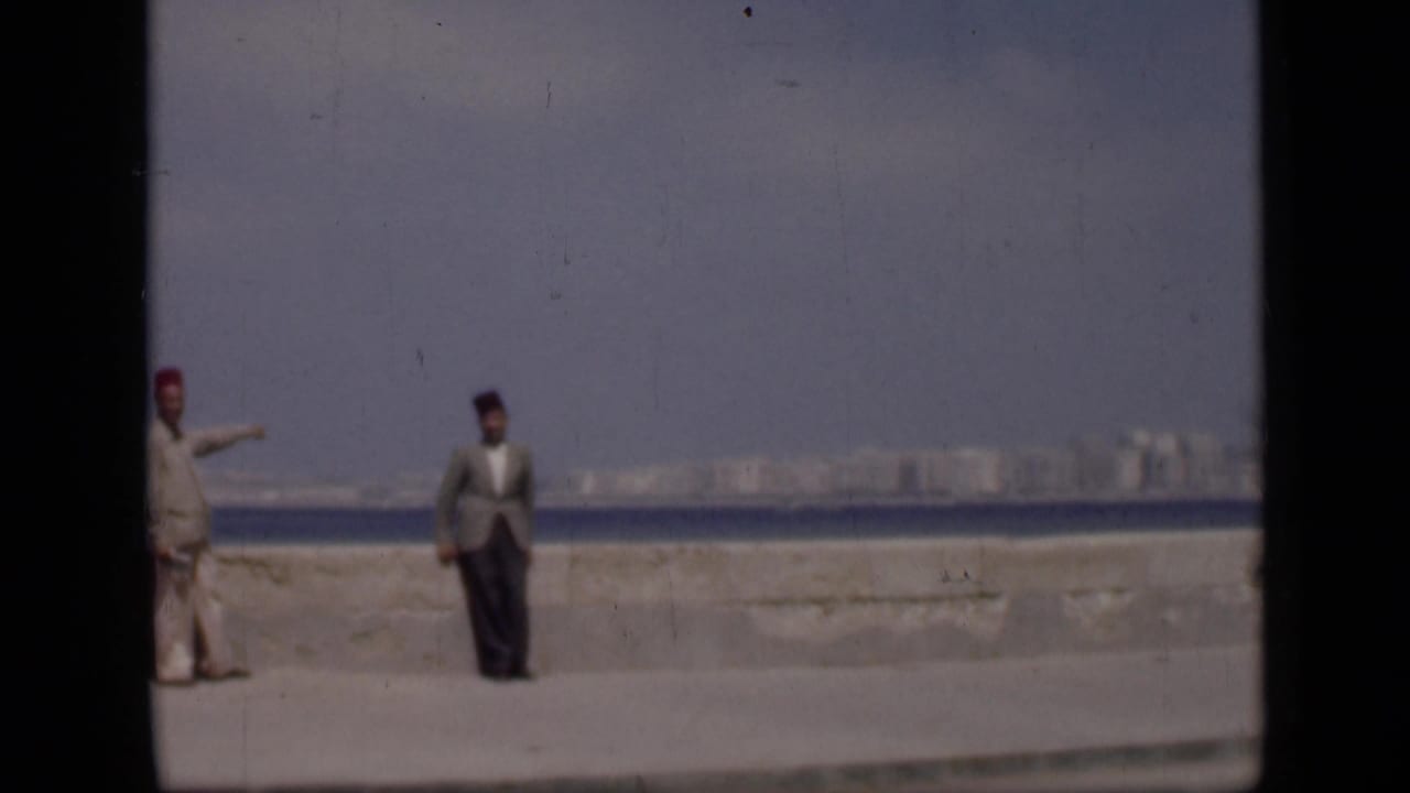 1946 Egypt home-movie frame showing two men in fez hats on a waterfront promenade with water and a distant skyline behind them.