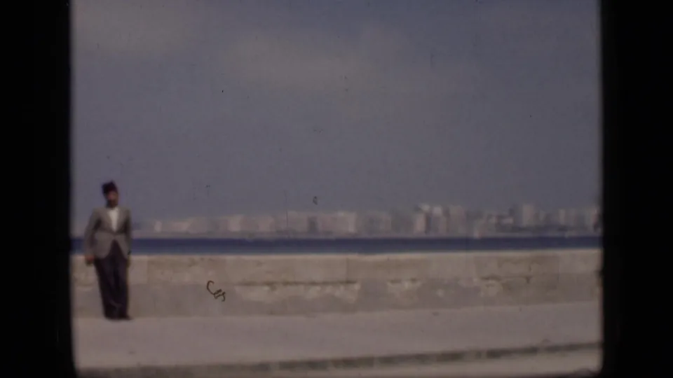 1946 Egypt home-movie frame with a suited man in a fez at the left edge, a stone seawall, open water, and distant buildings.