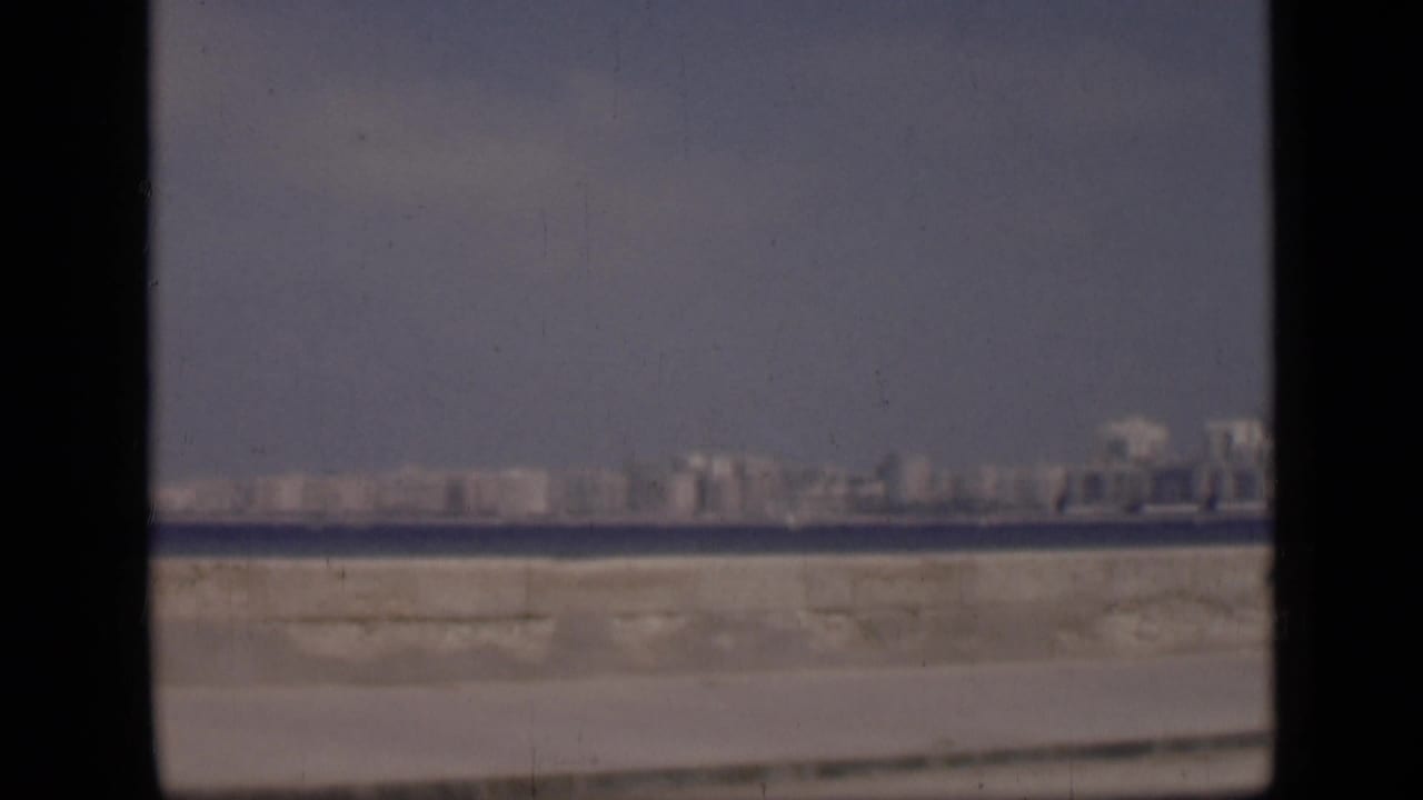 1946 Cairo home-movie frame showing a distant cityscape across water, with a hazy sky, pale embankment, and visible 8mm gate borders.