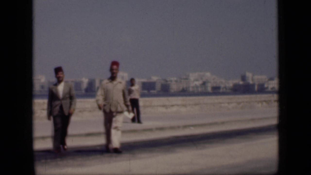 In 1946 Cairo, Egypt, two adult men in suits and fez hats walk toward the camera along a waterfront promenade, with another person and distant buildings visible.