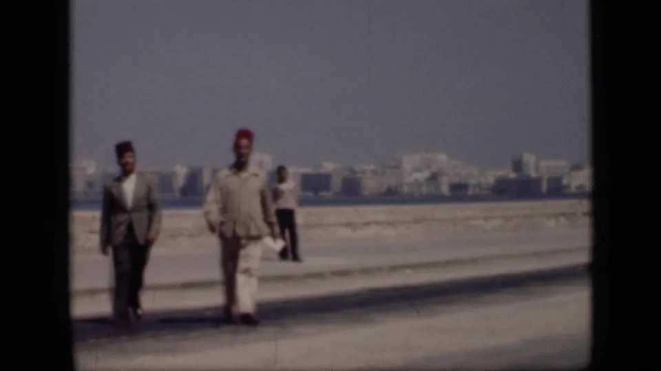 In 1946 Cairo, Egypt, two adult men in suits and fez hats walk toward the camera along a waterfront promenade, with another person and distant buildings visible.