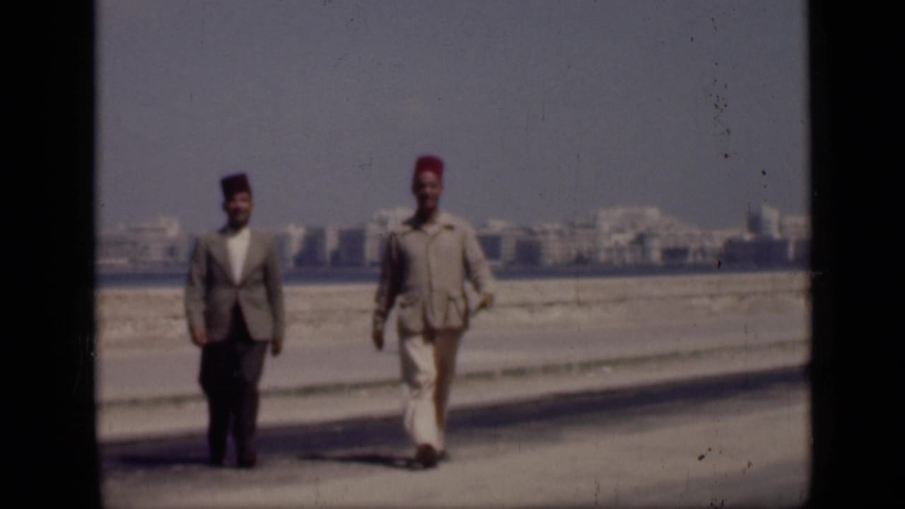 In 1946 Cairo, Egypt, two suited adult men wearing fez hats walk closer to the camera on a paved path beside open water and distant buildings.