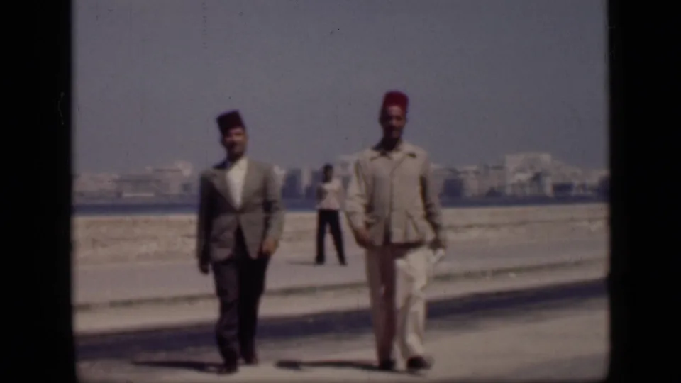 In 1946 Cairo, Egypt, the two men continue walking toward the camera on the promenade while a background figure stands near the waterfront.