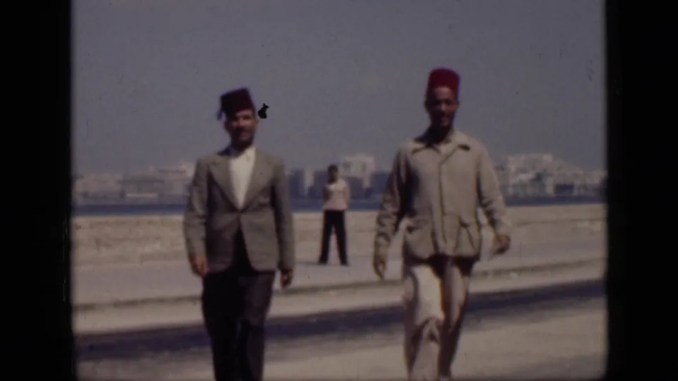 In 1946 Cairo, Egypt, two adult men in fez hats and suits pass close to the camera, with the waterfront and distant buildings soft in the background.