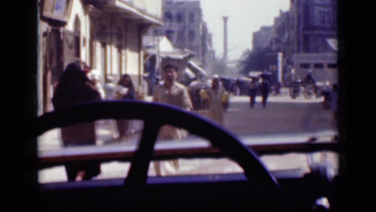 Street seen through a vehicle windshield with a steering wheel in front and pedestrians outside.