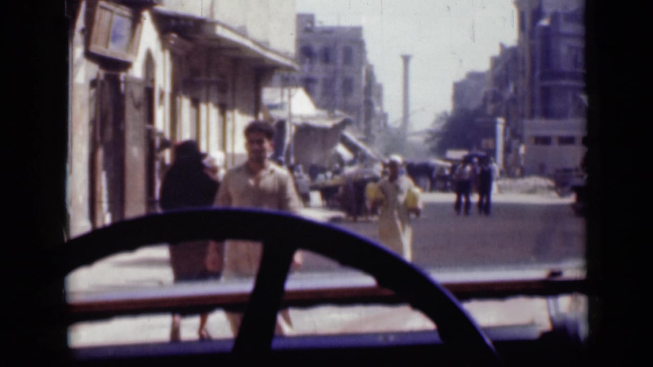 Vehicle POV of a Cairo street in 1946, showing pedestrians in light and dark clothing, shops, awnings, and taller buildings beyond.