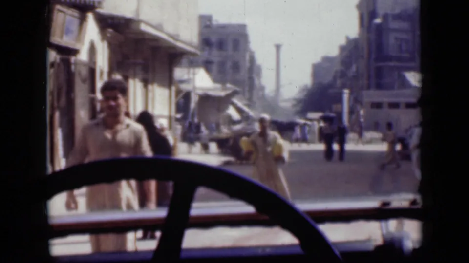 1946 Cairo vehicle-window view with a man close to the foreground, pedestrians farther back, shop awnings, and urban buildings beyond.