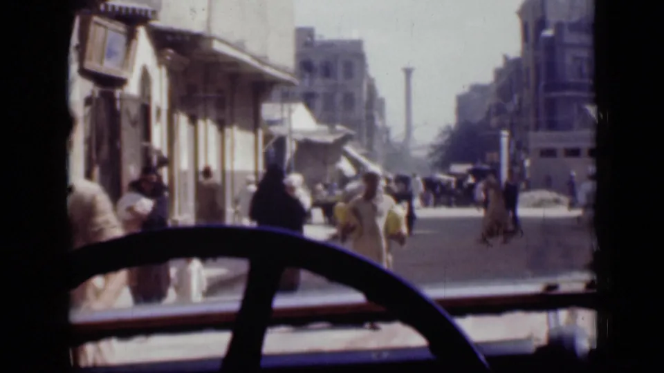 Street activity in Cairo, 1946, viewed from inside a vehicle with pedestrians, awnings, buildings, and a large steering wheel in the foreground.