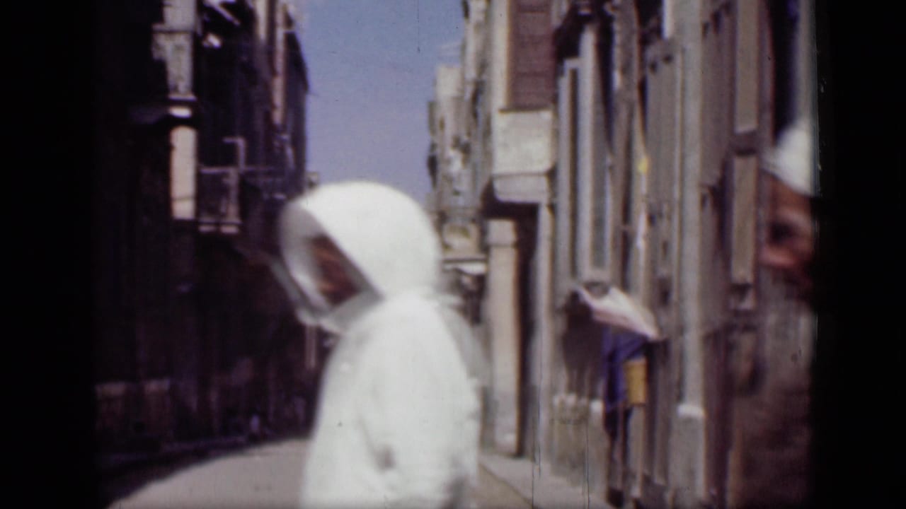 Pedestrian in a white head covering passes a narrow street with buildings on both sides.