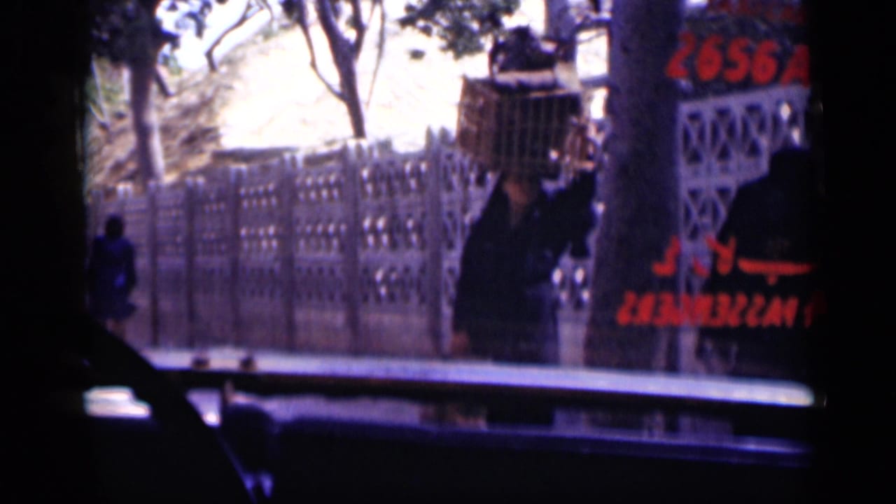 In Cairo archival home-movie footage, a woman carrying a large basket on her head passes a decorative fence and red Arabic script.