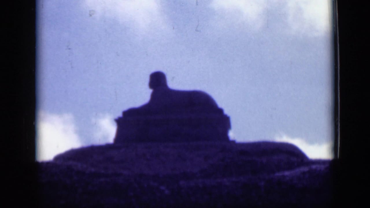 Dark silhouette of a large statue-like form on a raised hill against a pale cloudy sky.