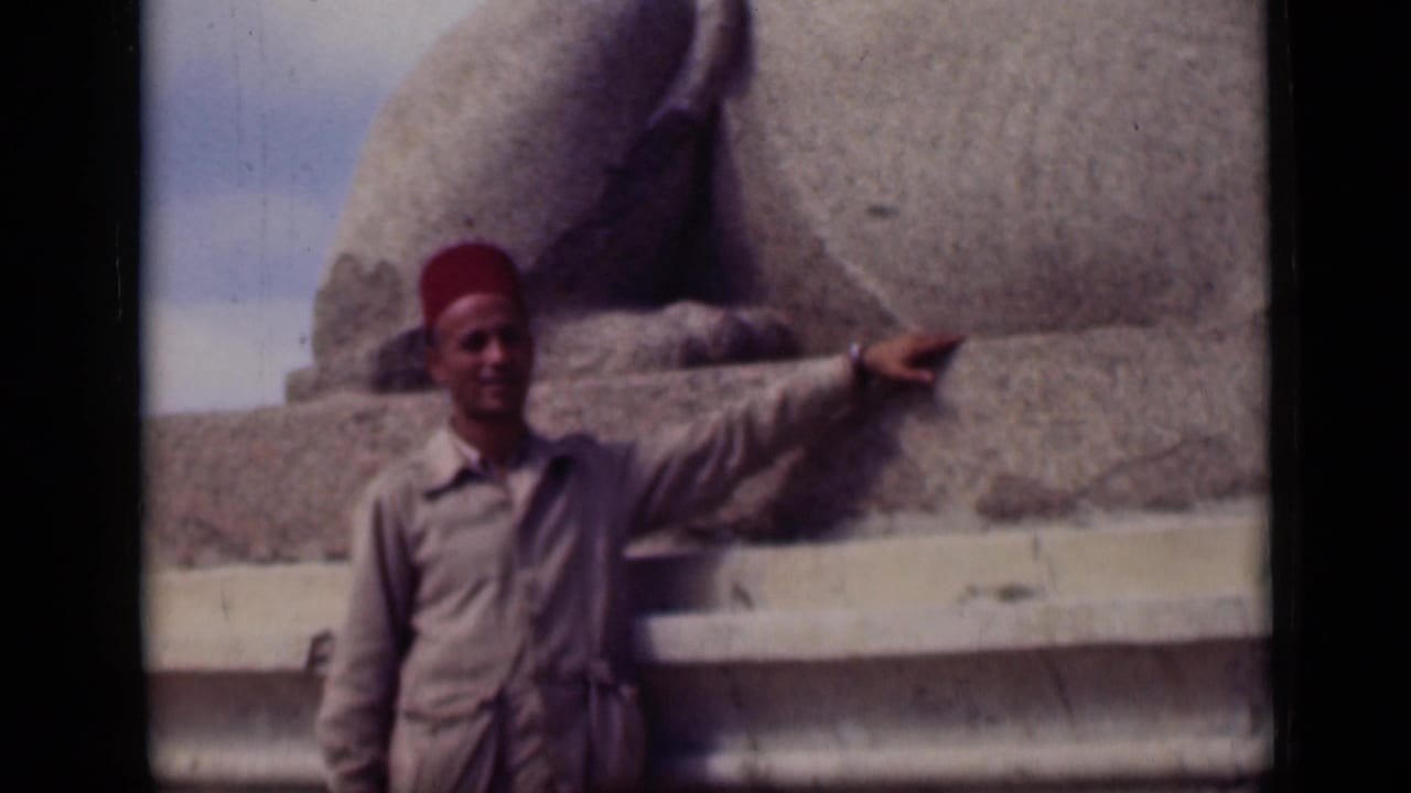 Adult man in a red fez and tan clothing stands at the Sphinx base in Cairo, extending one arm across the stonework.