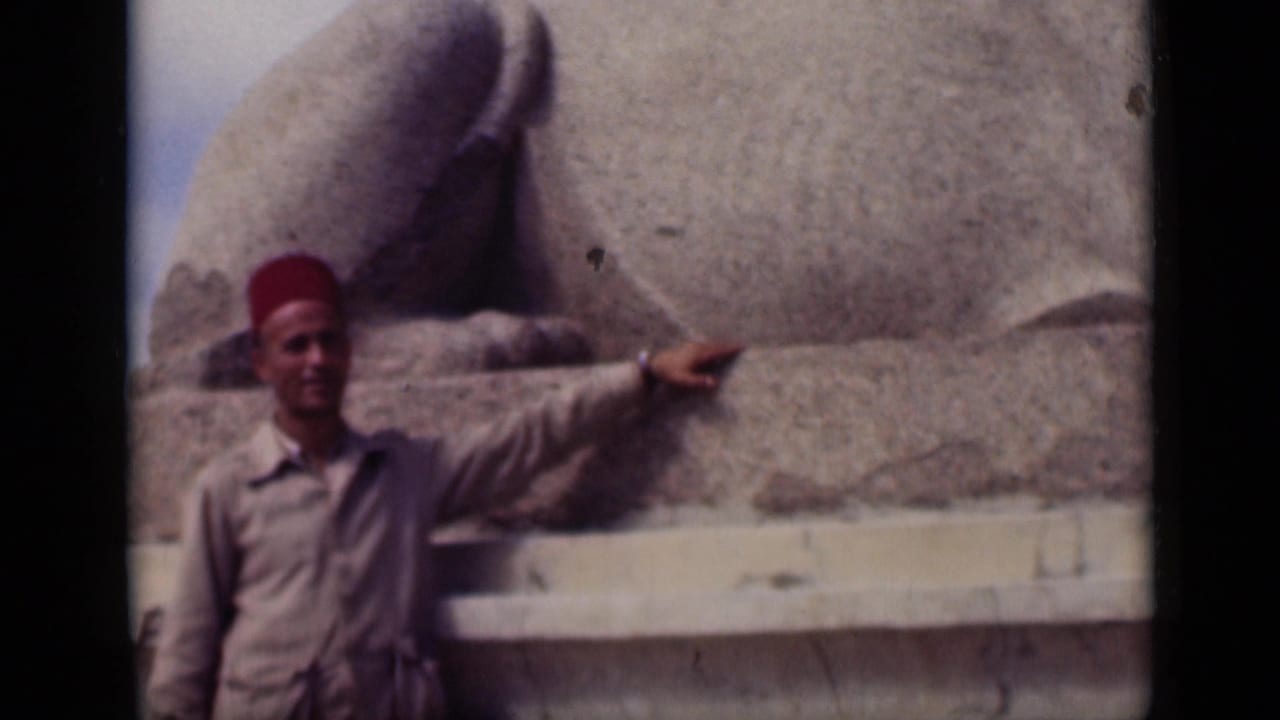 1946 Cairo home-movie still of a man in a red fez pointing beside the stone base of the Sphinx, with faded 8mm color.