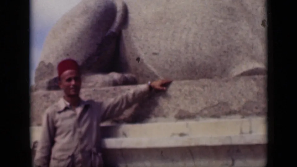 1946 Cairo home-movie still of a man in a red fez pointing beside the stone base of the Sphinx, with faded 8mm color.