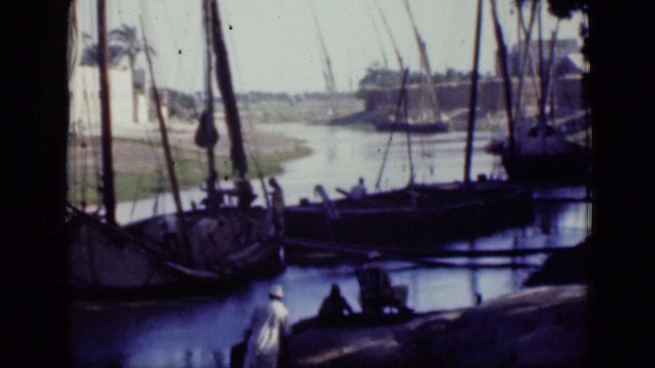 A man in light clothing walks along the shaded Cairo canal bank near docked wooden sailing boats and rigging.