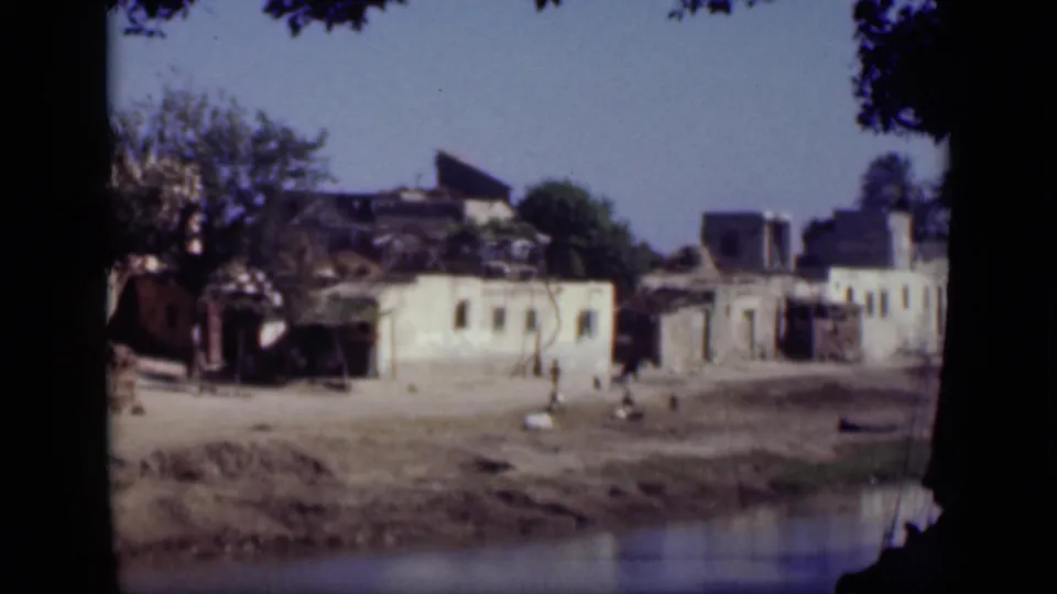 1946 Cairo footage frame with weathered low buildings, scattered people, and a calm water edge beneath dark tree framing.