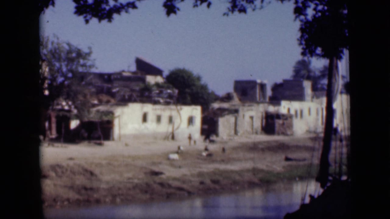 1946 Cairo home-movie frame showing damaged plaster buildings, a waterfront bank, tree shadows, and distant people crossing the open area.