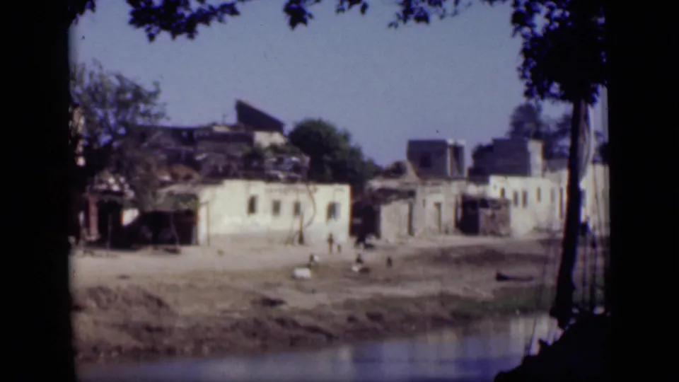 1946 Cairo home-movie frame showing damaged plaster buildings, a waterfront bank, tree shadows, and distant people crossing the open area.