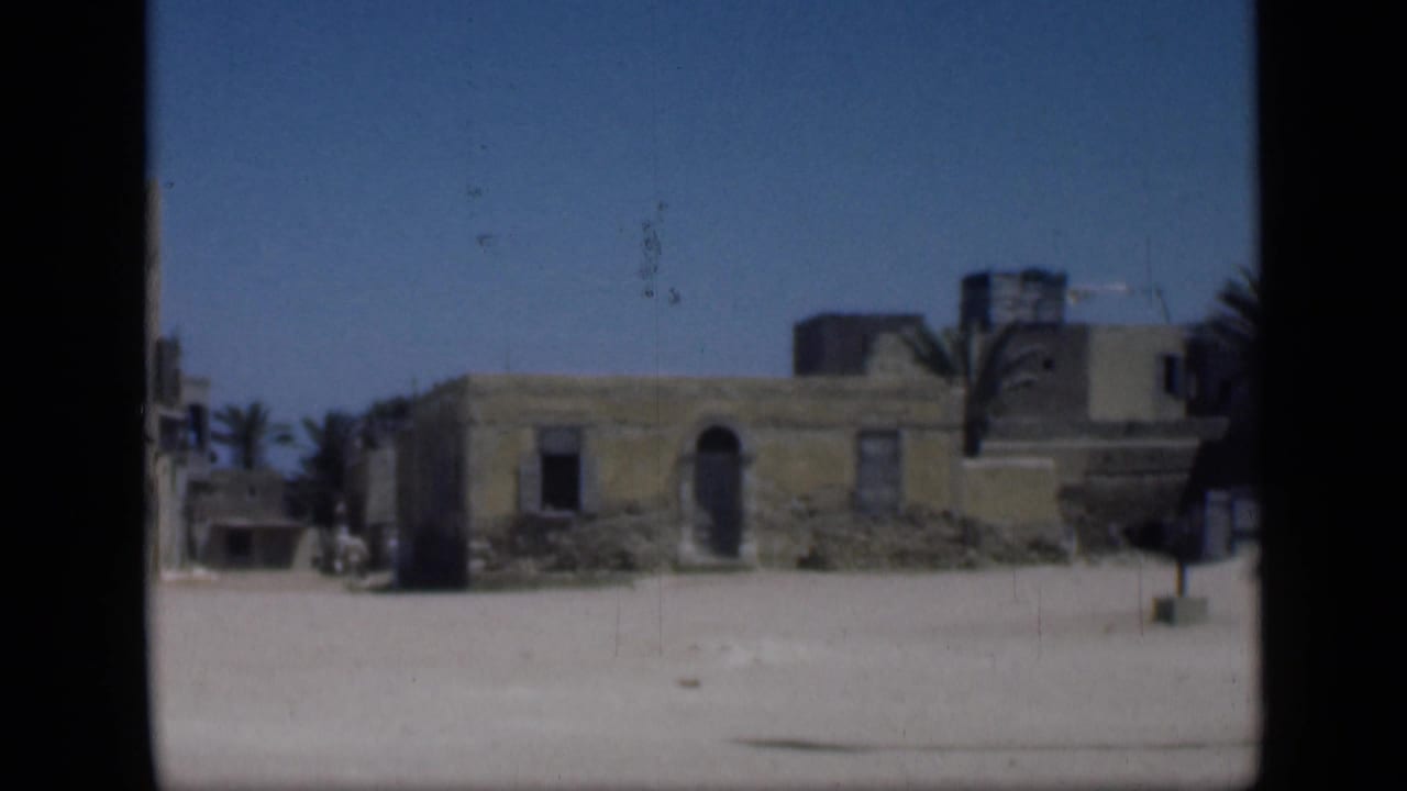 1946 Cairo-area 8mm frame showing low sand-colored buildings, dark window openings, palms, blue sky, and an empty-looking sandy foreground.
