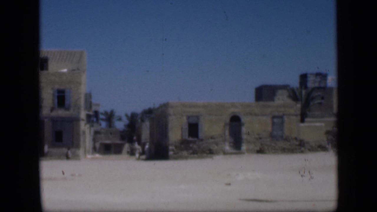Low weathered buildings, palms, and a sandy open area under blue sky.