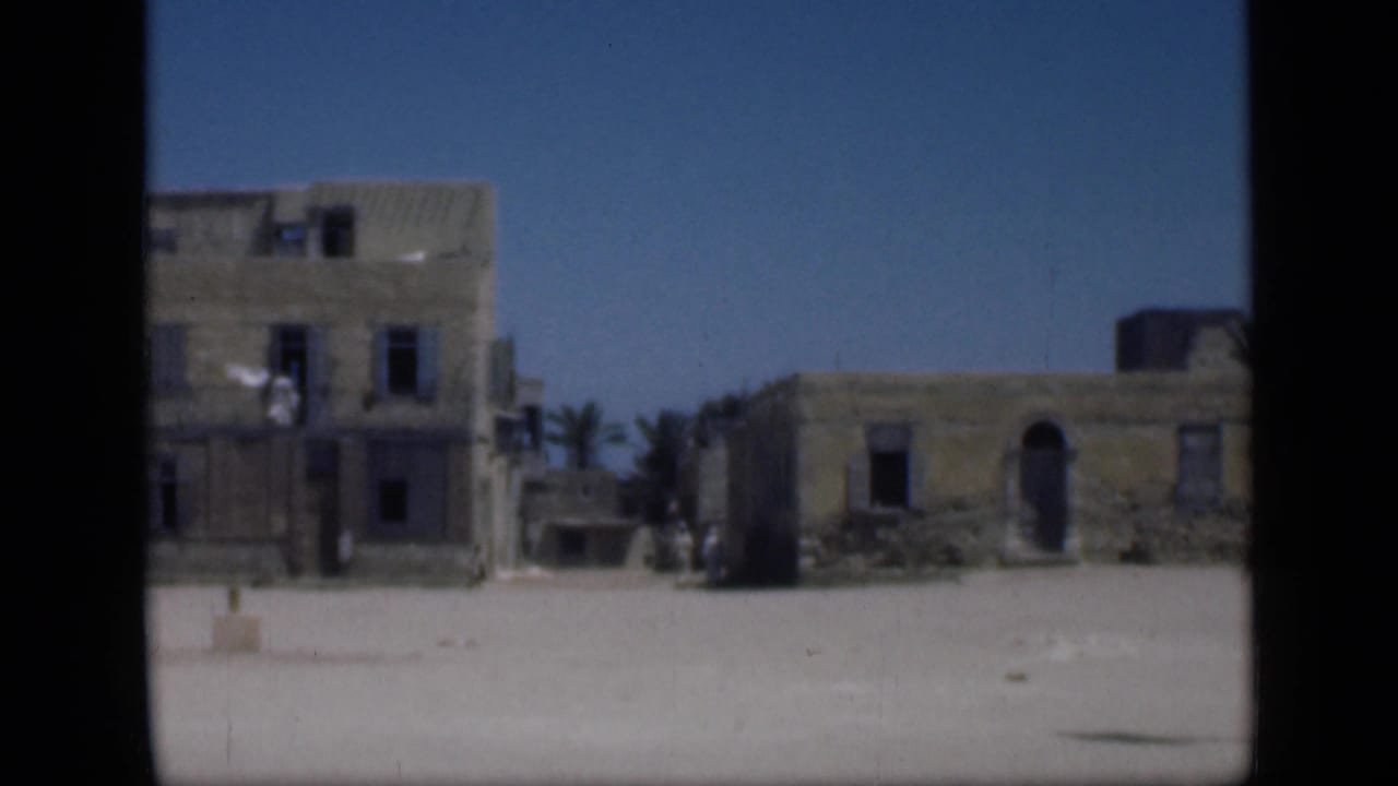 1946 Cairo-area 8mm frame showing a taller weathered building, adjacent low structures, palms, sandy ground, and possible tiny distant figures.