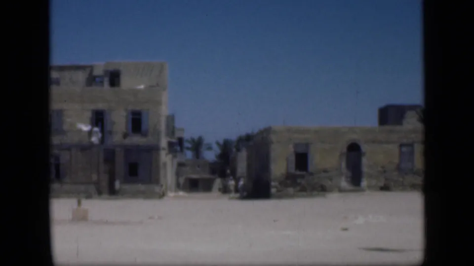 1946 Cairo-area 8mm frame showing a taller weathered building, adjacent low structures, palms, sandy ground, and possible tiny distant figures.