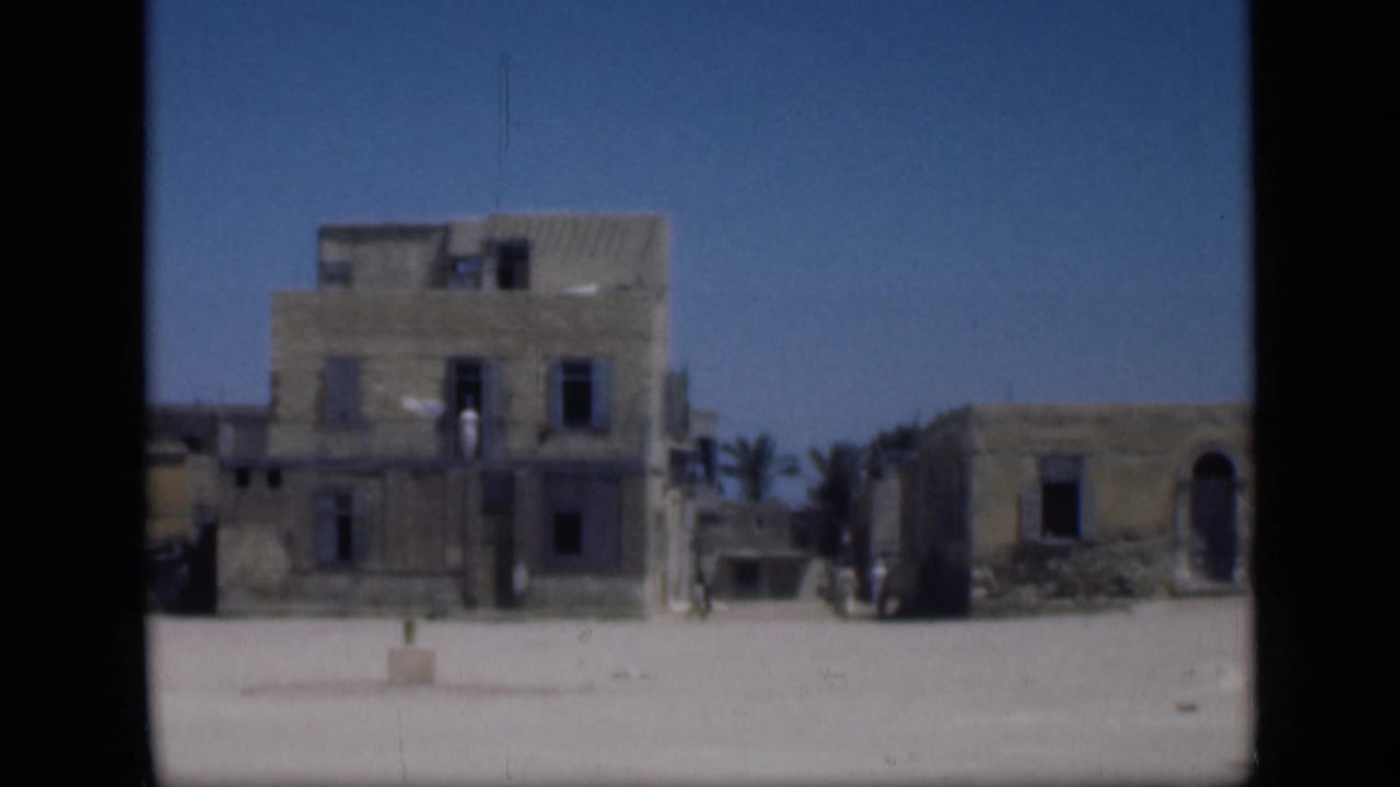 1946 Cairo-area frame with a taller stucco-like building, a lower arched structure, palms, open sandy ground, and small indistinct people in the distance.