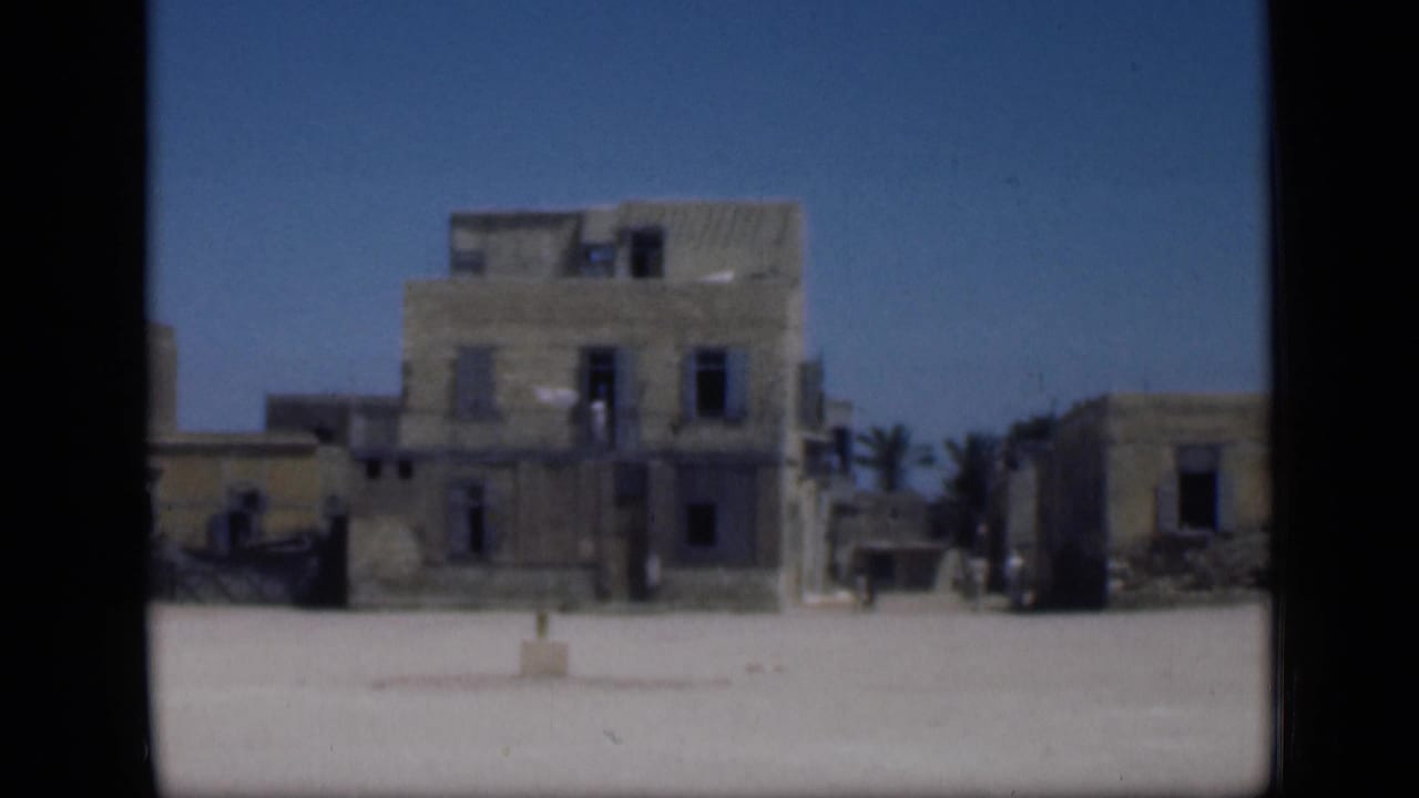 1946 Cairo home-movie frame showing weathered multi-story buildings across a bright open square, with dark windows and a clear blue sky.