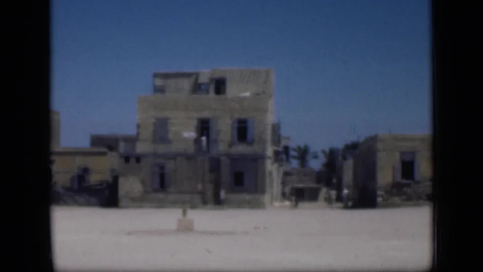 1946 Cairo home-movie frame showing weathered multi-story buildings across a bright open square, with dark windows and a clear blue sky.