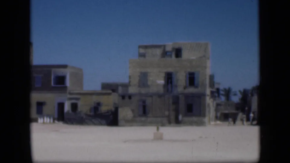 1946 Cairo exterior with old stucco or stone buildings, open ground, blue sky, and a distant pedestrian near the right side.