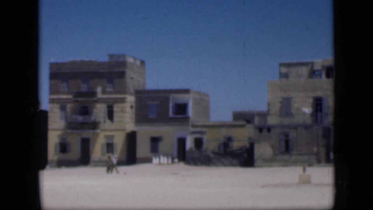 1946 Cairo home-movie frame with old multi-story buildings, open sunlit ground, and a distant pedestrian crossing the square.