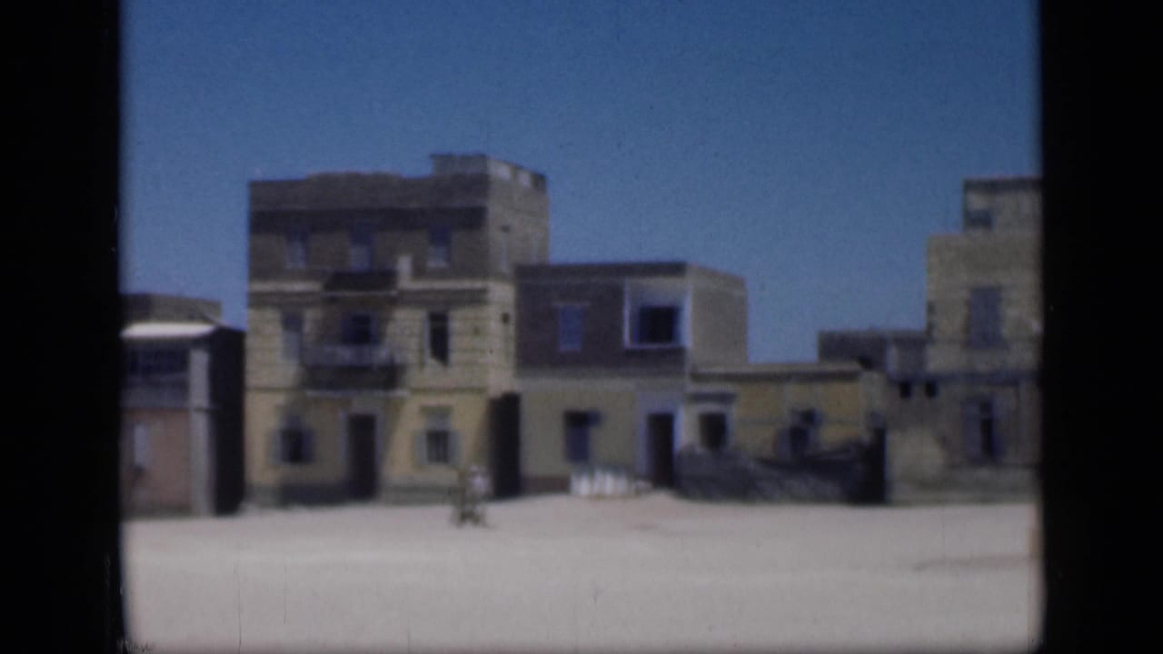 Sunlit flat-roofed buildings beside an open sandy foreground under a clear blue sky.