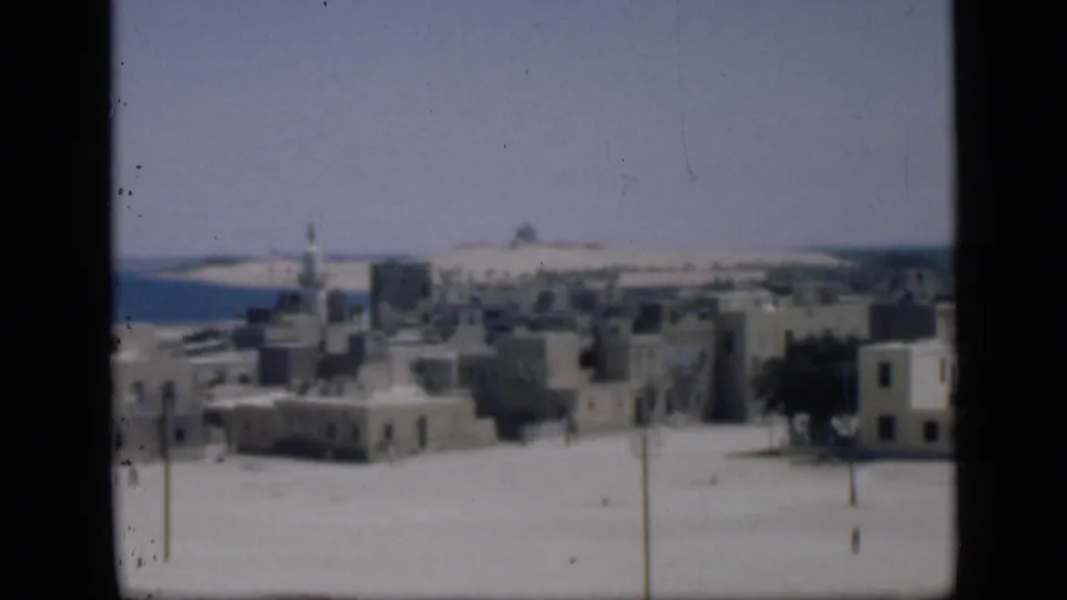 Final 8mm frame of the Cairo-listed clip showing a pale townscape, minaret, open sandy ground, distant horizon, and tree at right.