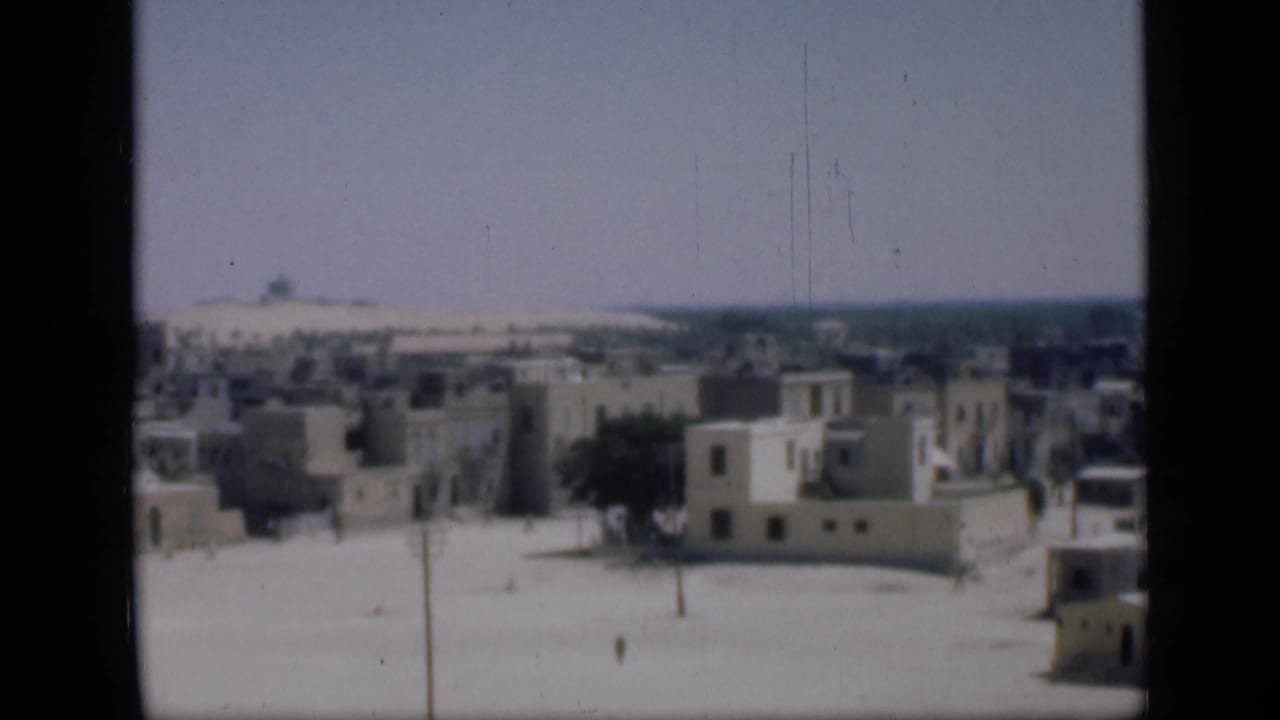 1946 Cairo home-movie frame showing pale flat-roofed buildings, trees, sandy open ground, poles, and a distant horizon.