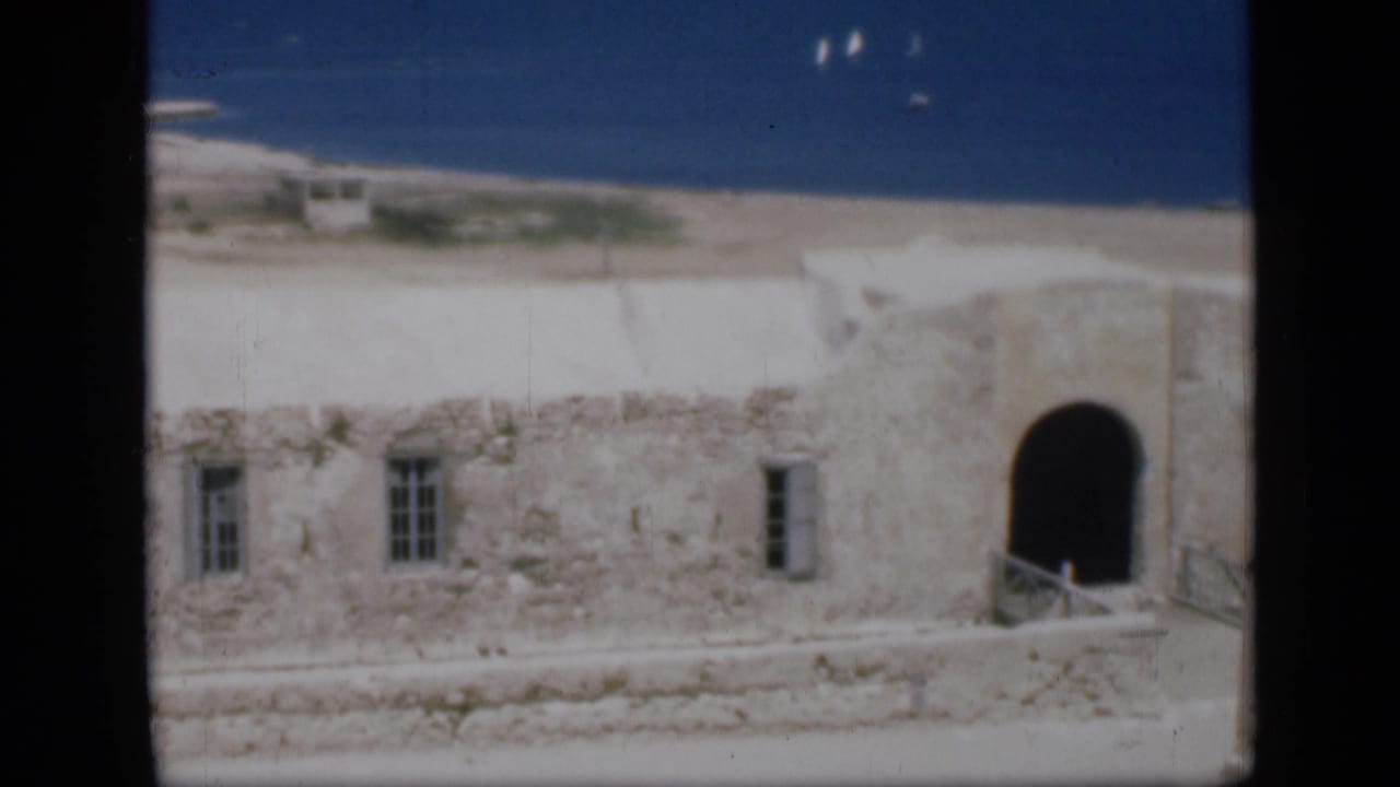 Whitewashed stone building beside a sandy beach and blue water.