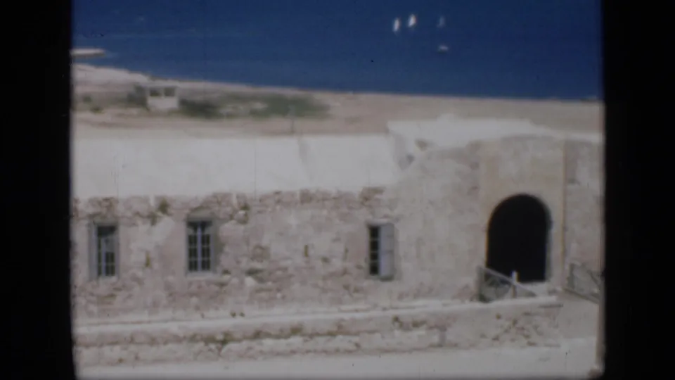 1946 Egypt home-movie frame showing a weathered white stone building with an arched doorway near sand and blue water.