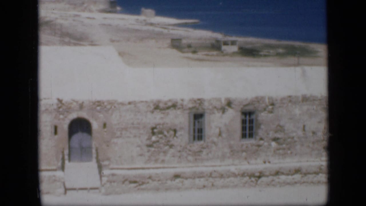 1946 Egypt frame showing an arched doorway, steps or walkway, barred windows, sand, and blue water beyond a white stone building.