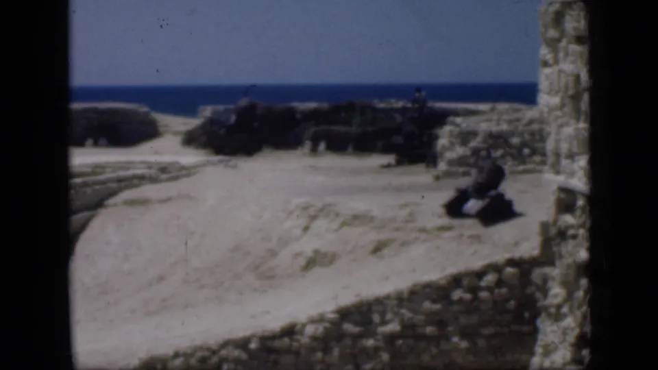 1946 Egypt 8mm frame showing sandy stone ruins, a vertical wall remnant, and blue water beyond the site.