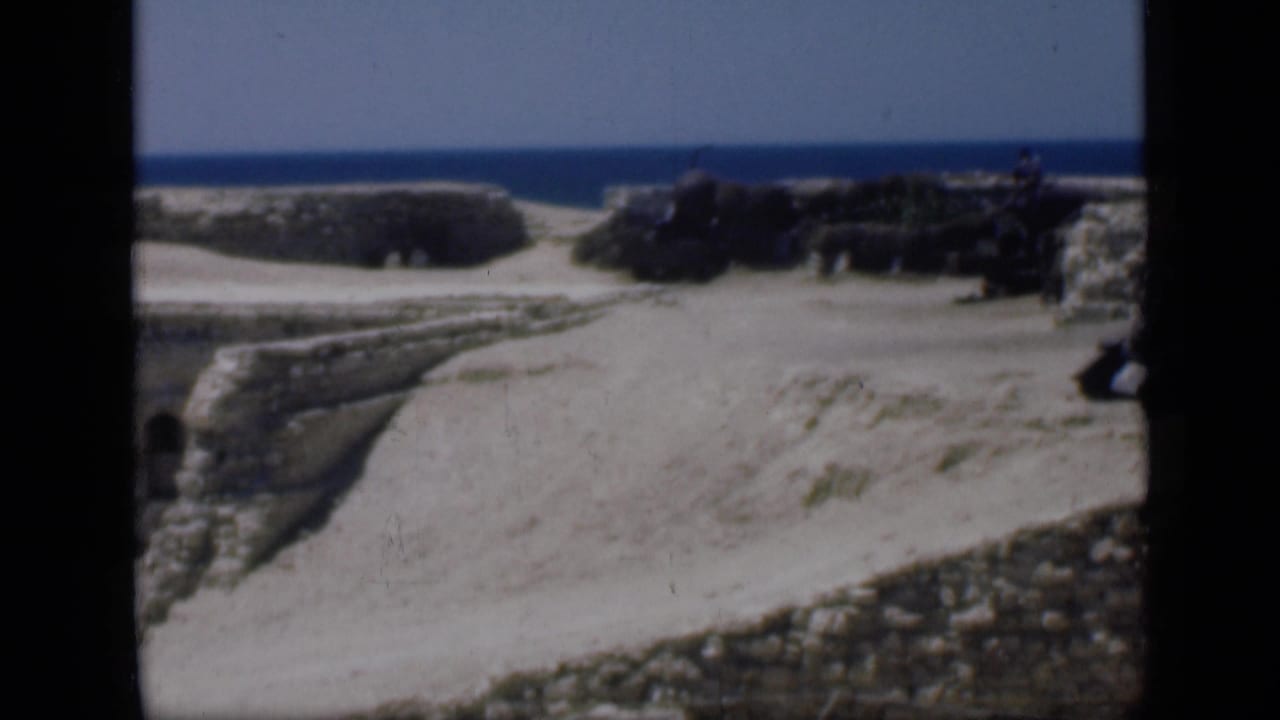 1946 Egypt travel footage frame with low stone ruins, pale sand, distant water, and a dark figure-like shape near the right edge.