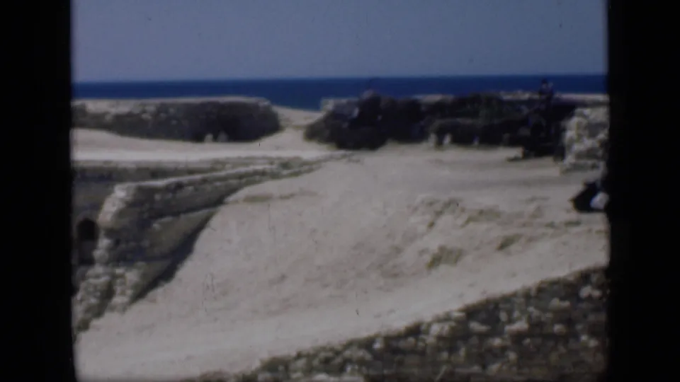 1946 Egypt travel footage frame with low stone ruins, pale sand, distant water, and a dark figure-like shape near the right edge.