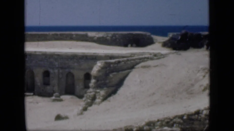 1946 Egypt frame showing a weathered stone structure with small arched openings, pale sand, and distant blue water.