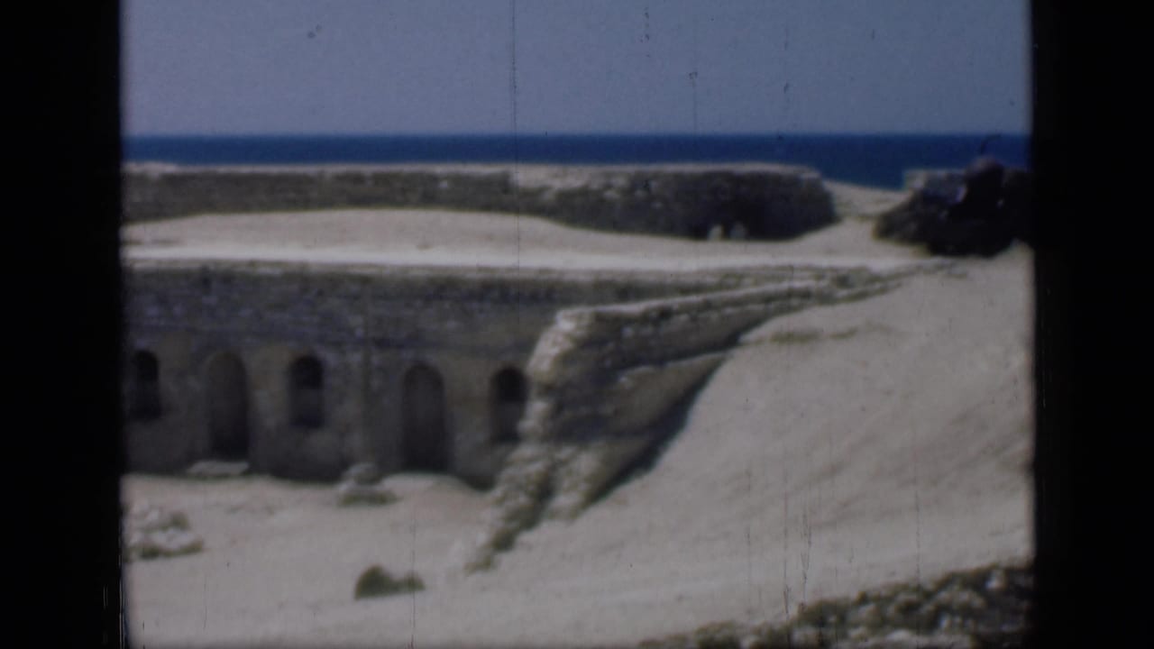 Stone fortification with arched doorways, sandy ground, and sea on the horizon.